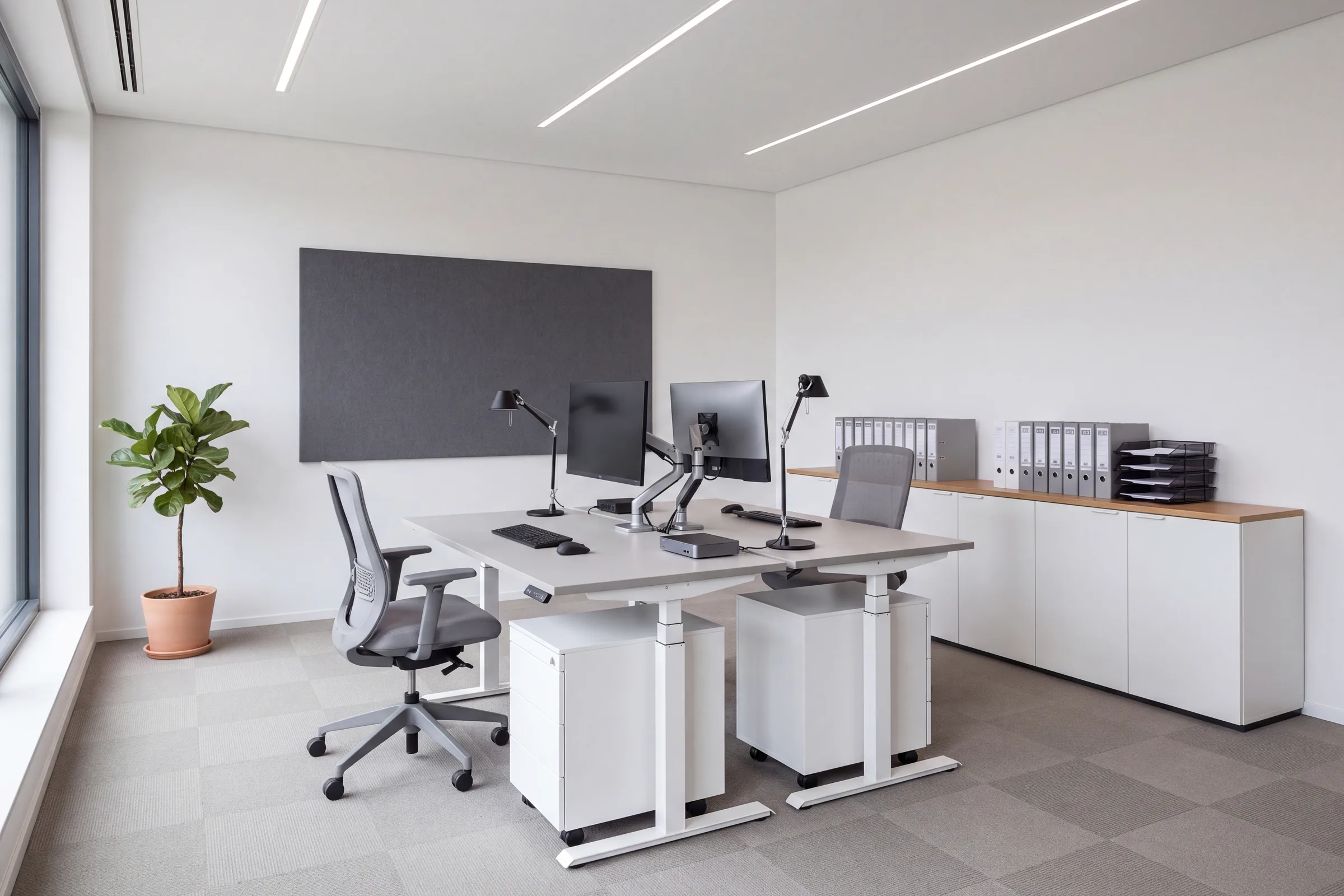 Zwei höhenverstellbare Schreibtische mit Monitoren und Bürostühlen, dahinter ein Aktenschrank mit Ordnern und Ablagekörben